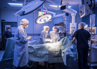 Four doctors in hospital operating room with patient lying on operating table. Surgical lights shining on medical team performing operation on patient