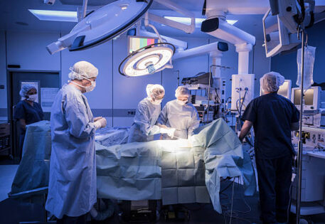 Four doctors in hospital operating room with patient lying on operating table. Surgical lights shining on medical team performing operation on patient